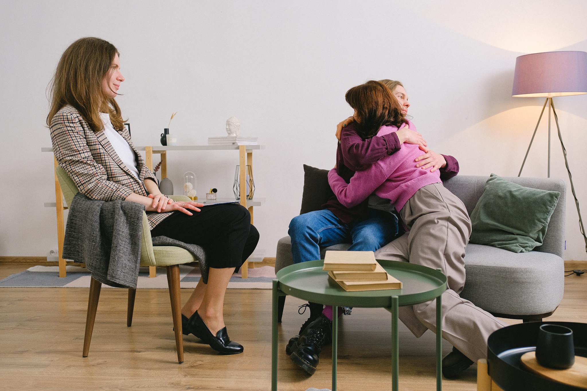 Side view of positive female therapist looking at loving couple cuddling on sofa near table with books